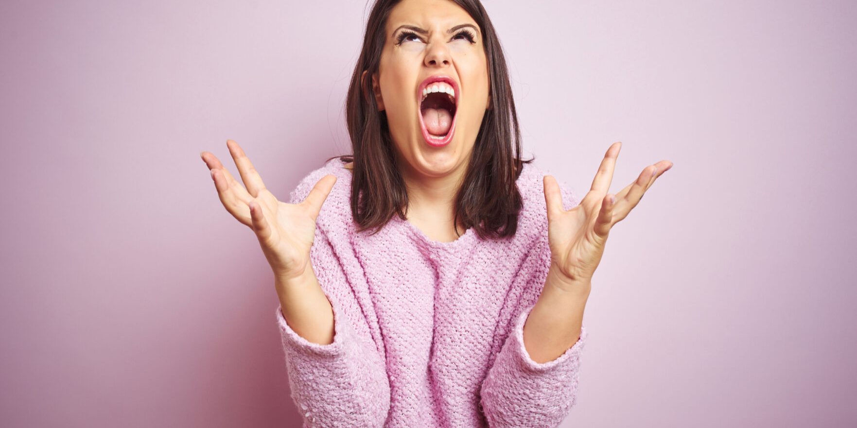 Young beautiful brunette woman wearing a sweater over pink isolated background crazy and mad shouting and yelling with aggressive expression and arms raised. Frustration concept.