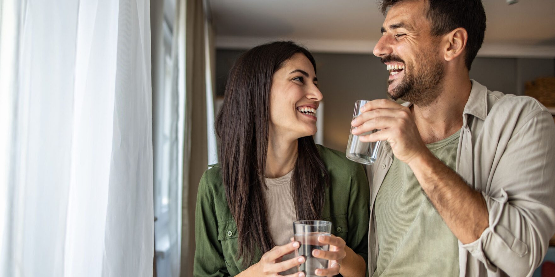 An adult couple enjoys a candid moment together by a window, exchanging gentle glances while holding glasses of water. Their deep connection and peaceful environment capture the essence of intimacy and mutual understanding. Ideal for themes of romance, emotional closeness and shared moments of joy.